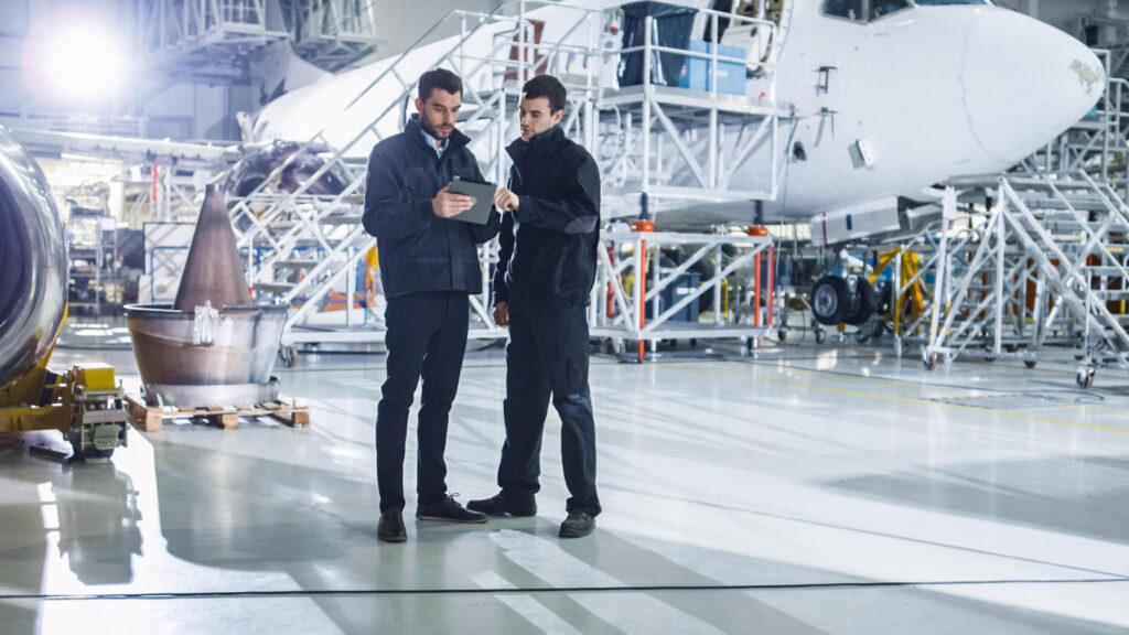 Ausbildung am Flughafen: Ausbilder und Auszubildender mit Tablet im Wartungshangar vor einem Flugzeug